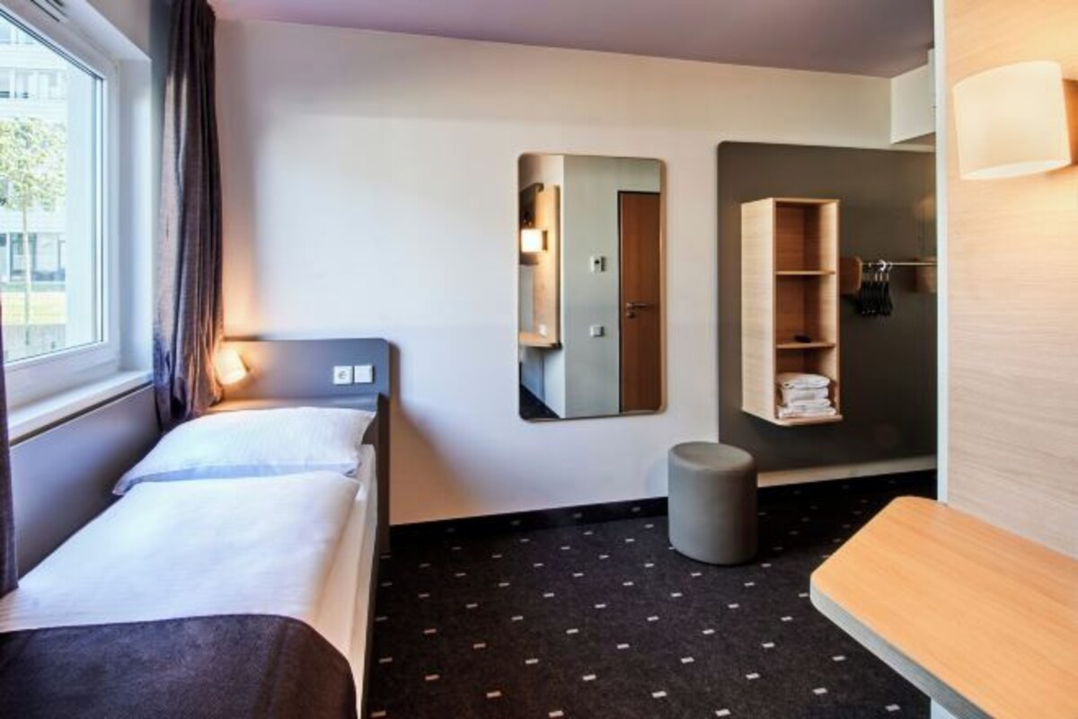 Ein Hotelzimmer mit einem Bett, einem kleinen Hocker, einem Spiegel und einem Regal mit Handtüchern.