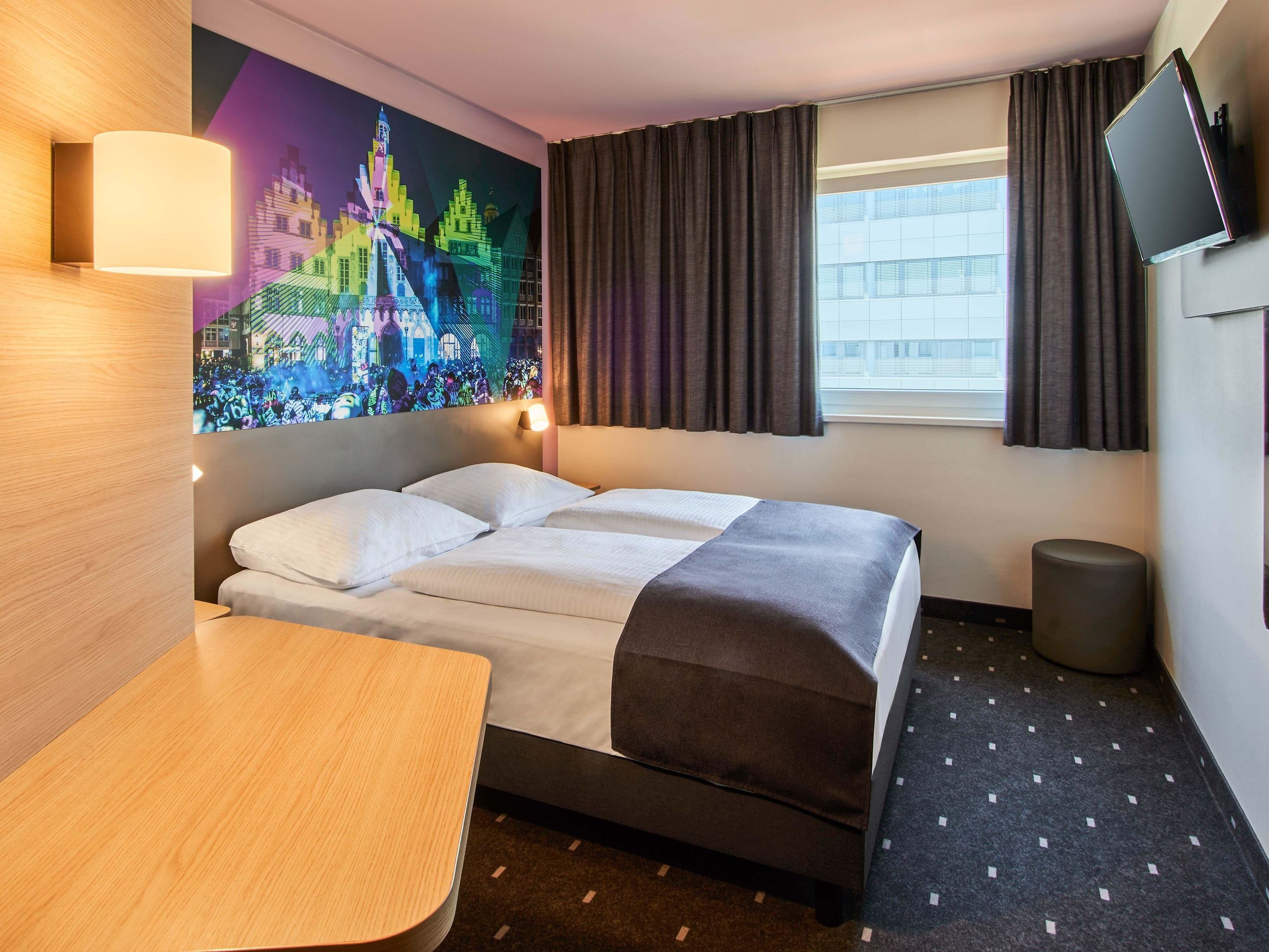 Ein Hotelzimmer mit Bett, Schreibtisch, Wand-Fernseher, einem Fenster mit Vorhängen und einem farbenfrohen Wandgemälde.