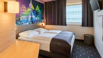 Ein Hotelzimmer mit Bett, Schreibtisch, Wand-Fernseher, einem Fenster mit Vorhängen und einem farbenfrohen Wandgemälde.