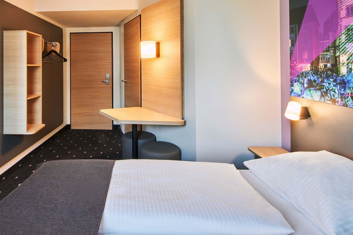 A hotel room with a bed, a desk, a chair, a wardrobe, and a mirror.