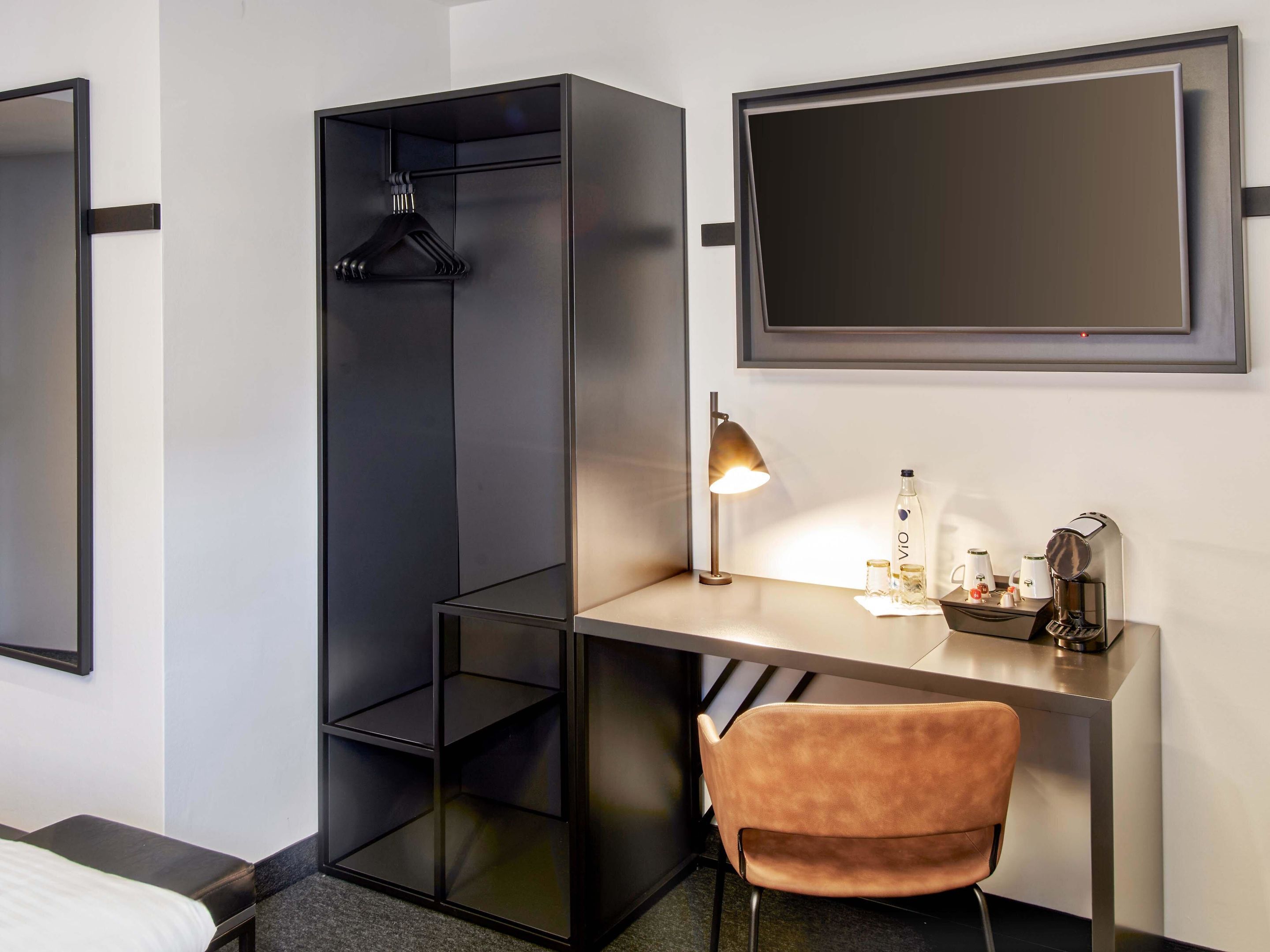 Una habitación de hotel moderna con escritorio, una silla, perchero, mesita de noche y un espejo grande.