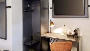 A modern hotel room with a desk, a chair, a hanging coat rack, a bedside table, and a large mirror.