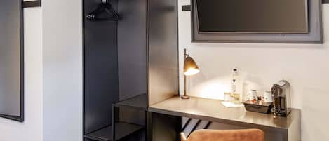 A modern hotel room with a desk, a chair, a hanging coat rack, a bedside table, and a large mirror.