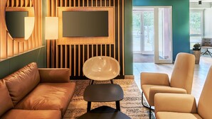 A modern living room with a sofa, armchair, and a coffee table. A flat-screen TV is mounted on the wall, and there is a large window providing natural light. - B&B Hotel Frankfurt-Niederrad (Frankfurt)