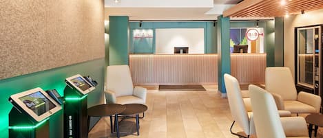 Um hall de hotel com balcão de recepção, totens de auto-check-in, área de estar e design moderno com detalhes em madeira.