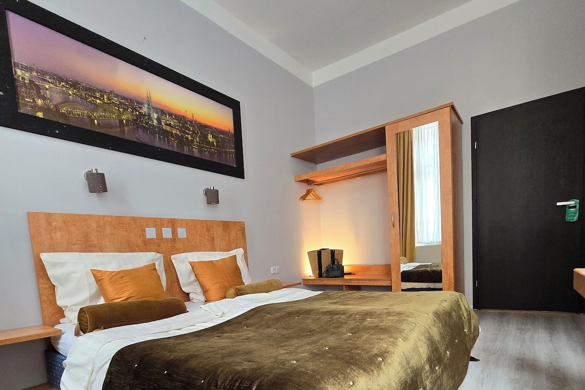 A hotel room with a bed, a nightstand, a wooden wardrobe, a framed cityscape picture, and a window with curtains.