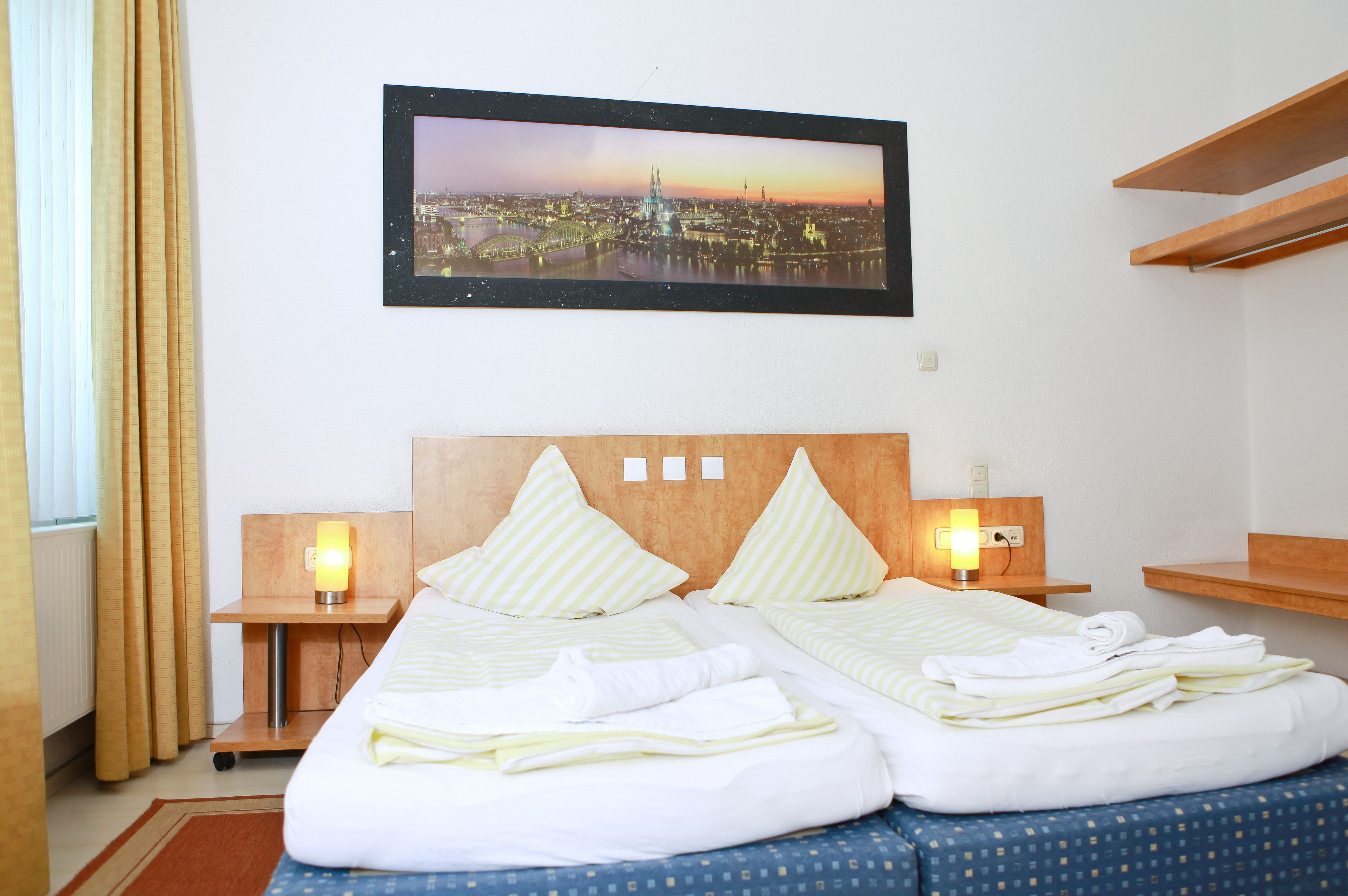 A hotel room with a bed, two bedside tables with lamps, a framed cityscape picture on the wall, and a window with curtains.