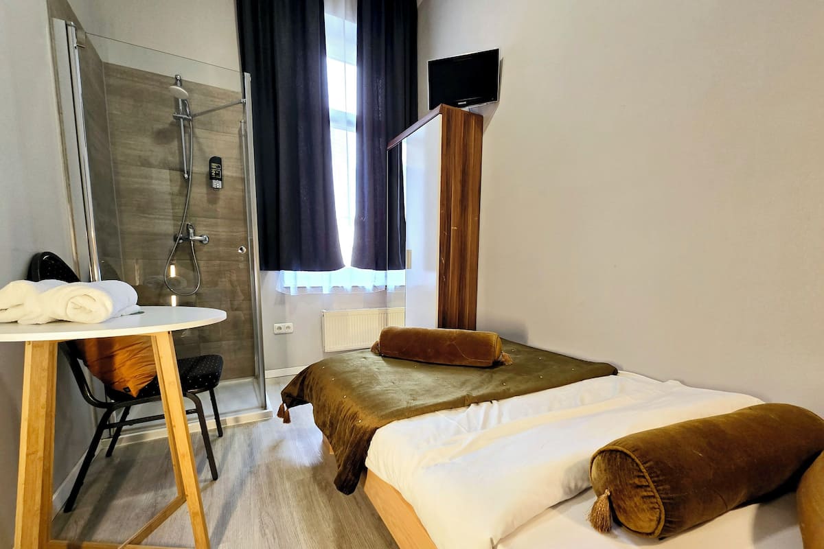 A hotel room with a bed, a desk with a chair, a shower area, and a television mounted on the wall.