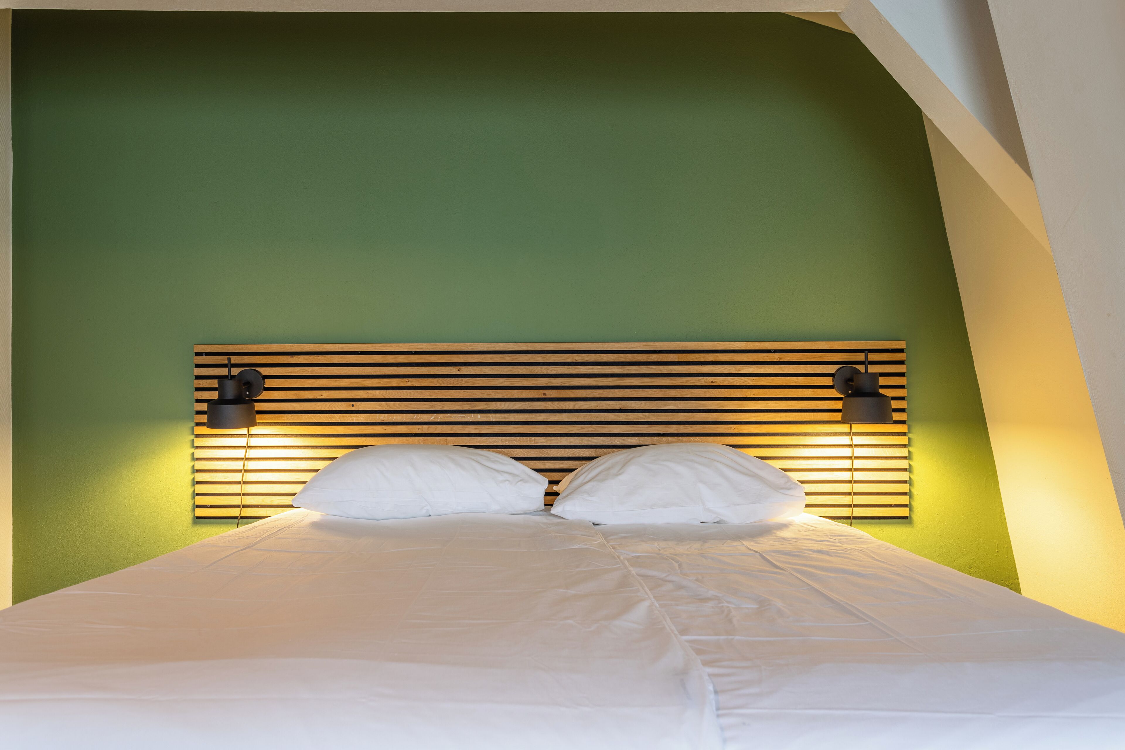 A bed with white linens, a wooden headboard, and two wall-mounted lamps.