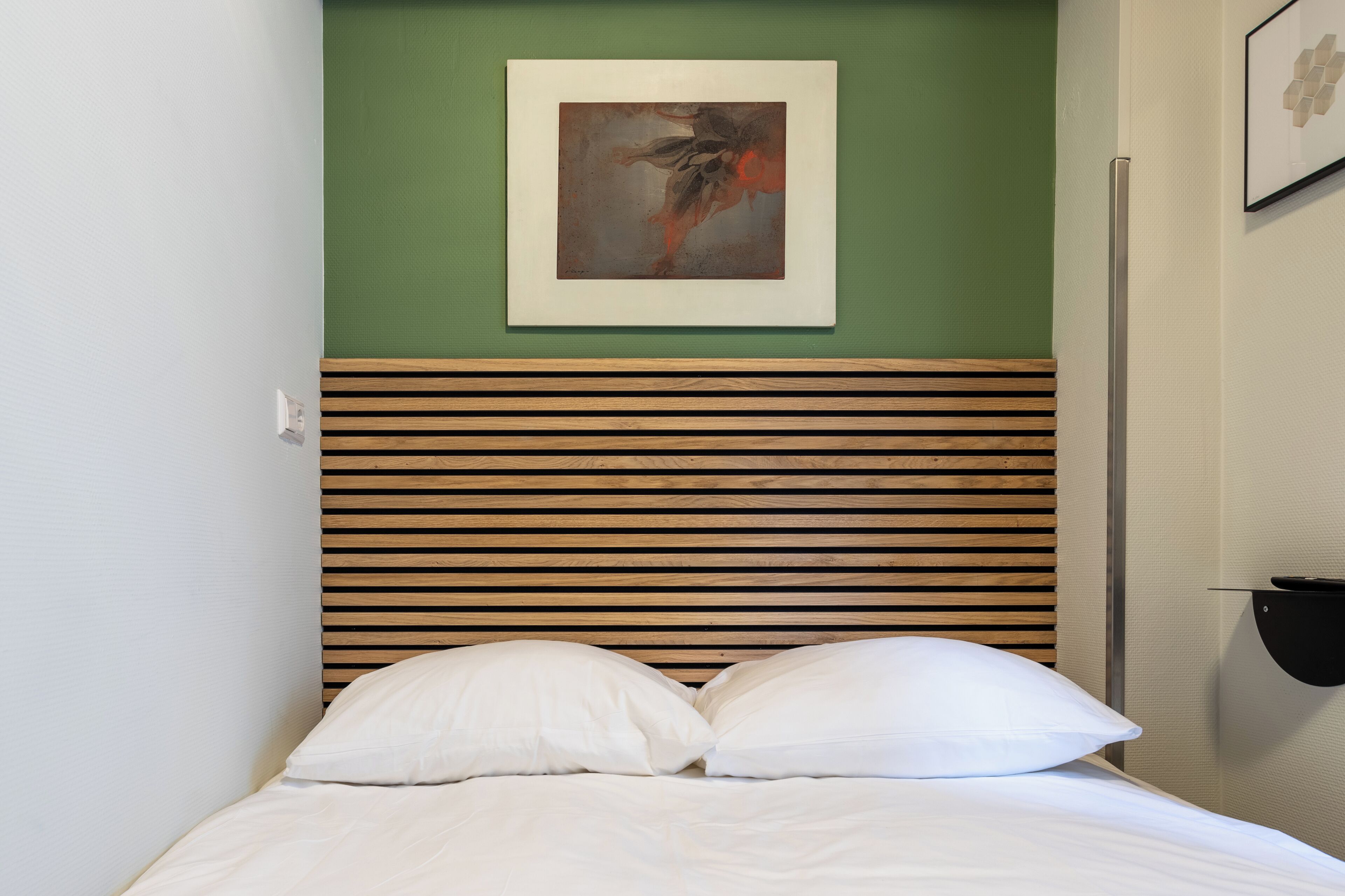 Een net opgemaakt bed met witte lakens en kussens, een houten lattenbovenstuk en een abstract schilderij in een lijst aan de muur.