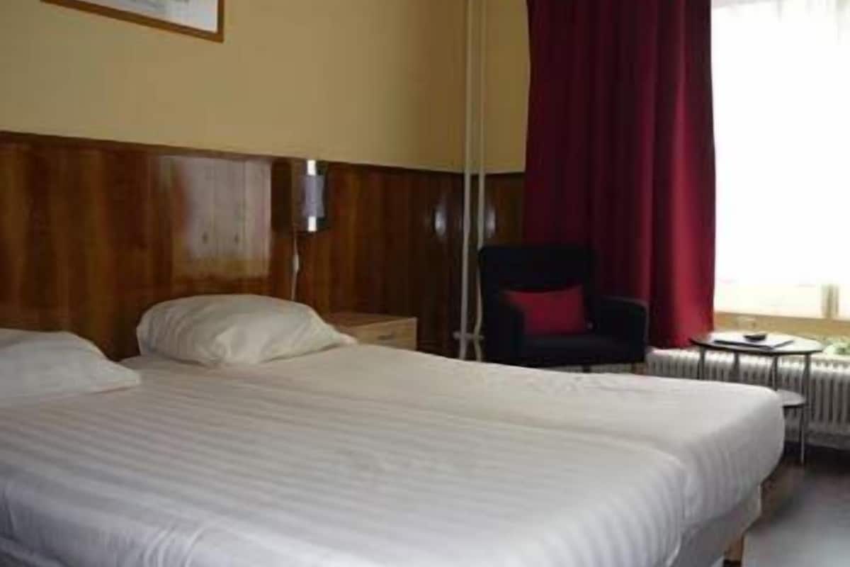 A hotel room with a large bed, a chair, a small table, and a window with curtains.
