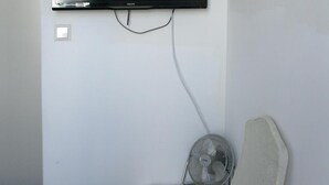 A room with a mounted flat-screen TV, a white chair, a small table with a remote control, and a standing fan. - Hotel Kap (Amsterdam)