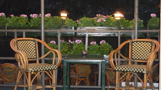 A table and two wicker chairs with checkered cushions, set against a backdrop of green foliage and pink flowers, under a metal railing. - Hotel Kap (Amsterdam)