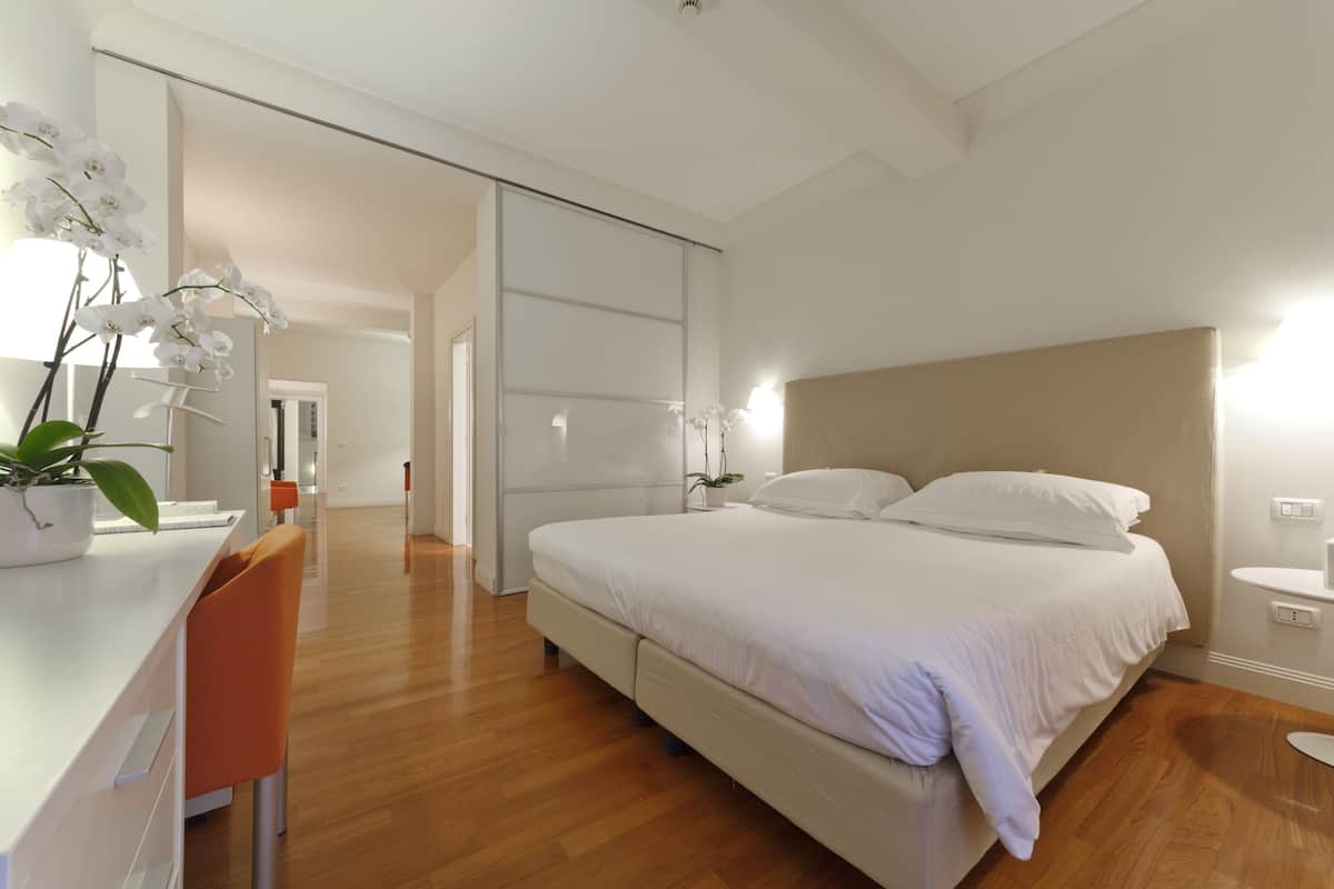 A modern bedroom with a large bed, a desk, and a chair. There is a potted plant on the desk and a mirror on the wall.