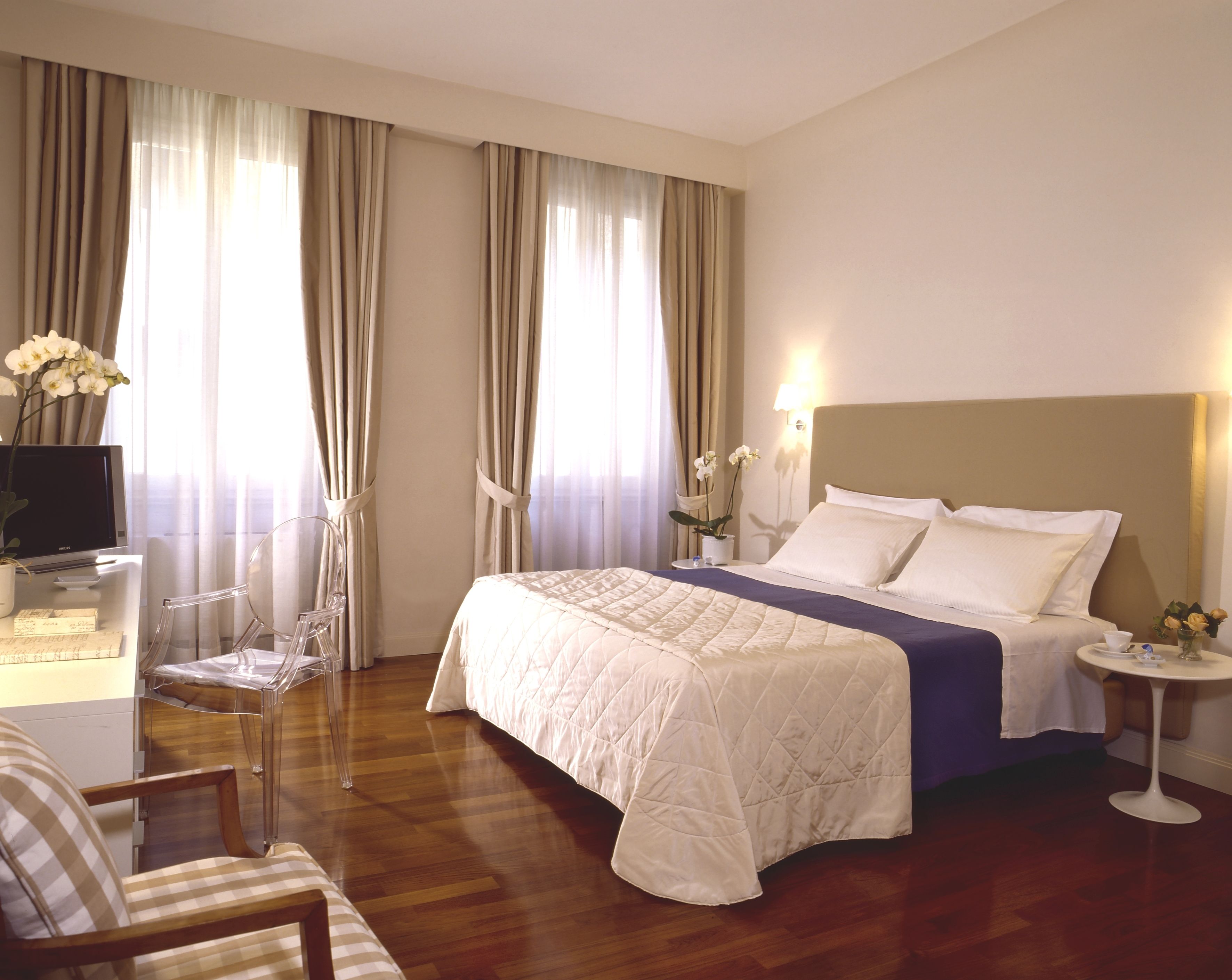 Habitación de hotel con cama, escritorio, computadora, silla y una pequeña mesa con un jarrón de flores.