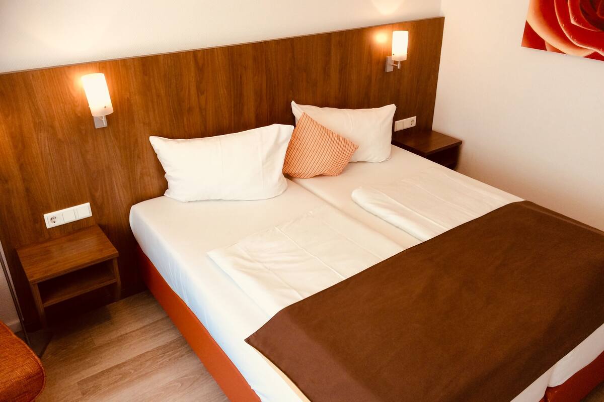 Cama bem arrumada com roupa de cama branca, manta marrom e duas almofadas, uma branca e outra laranja. Há duas mesas de cabeceira com abajures, e o quarto tem painéis de madeira nas paredes.