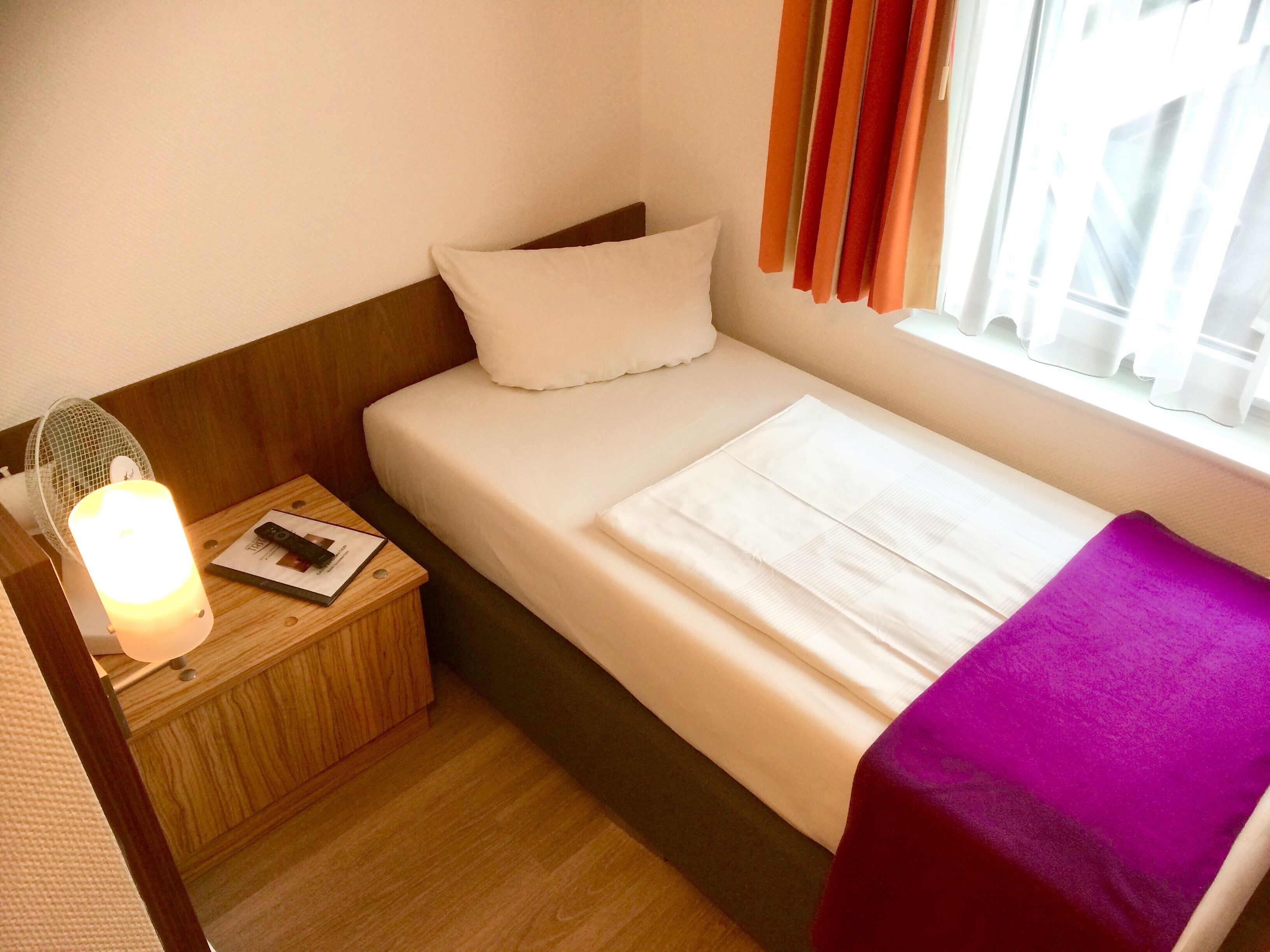 Ein kleines, ordentlich bezogenes Bett mit einem weißen Kopfkissen und einer violetten Decke, ein Nachttisch aus Holz mit einer Lampe sowie ein Fenster mit orangefarbenen Vorhängen.