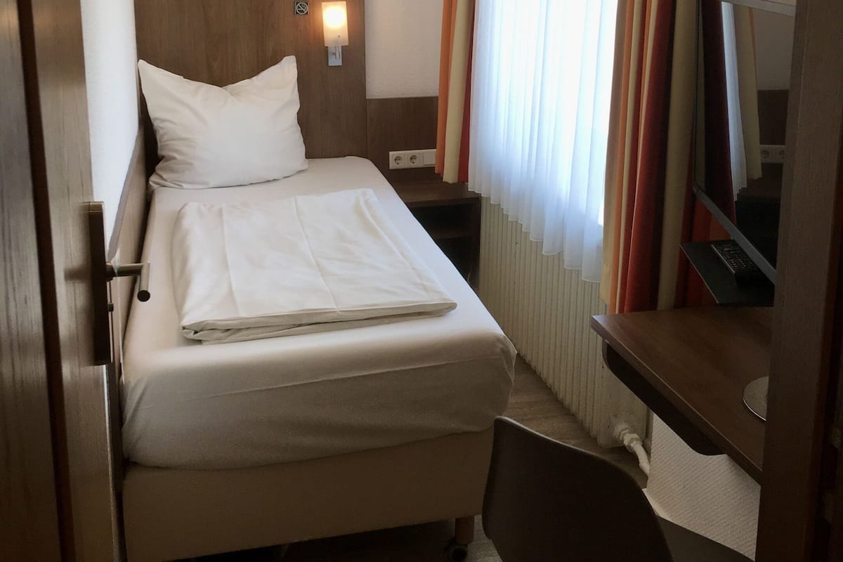 Quarto de hotel com cama, escrivaninha, cadeira, televisão e janela com cortinas.