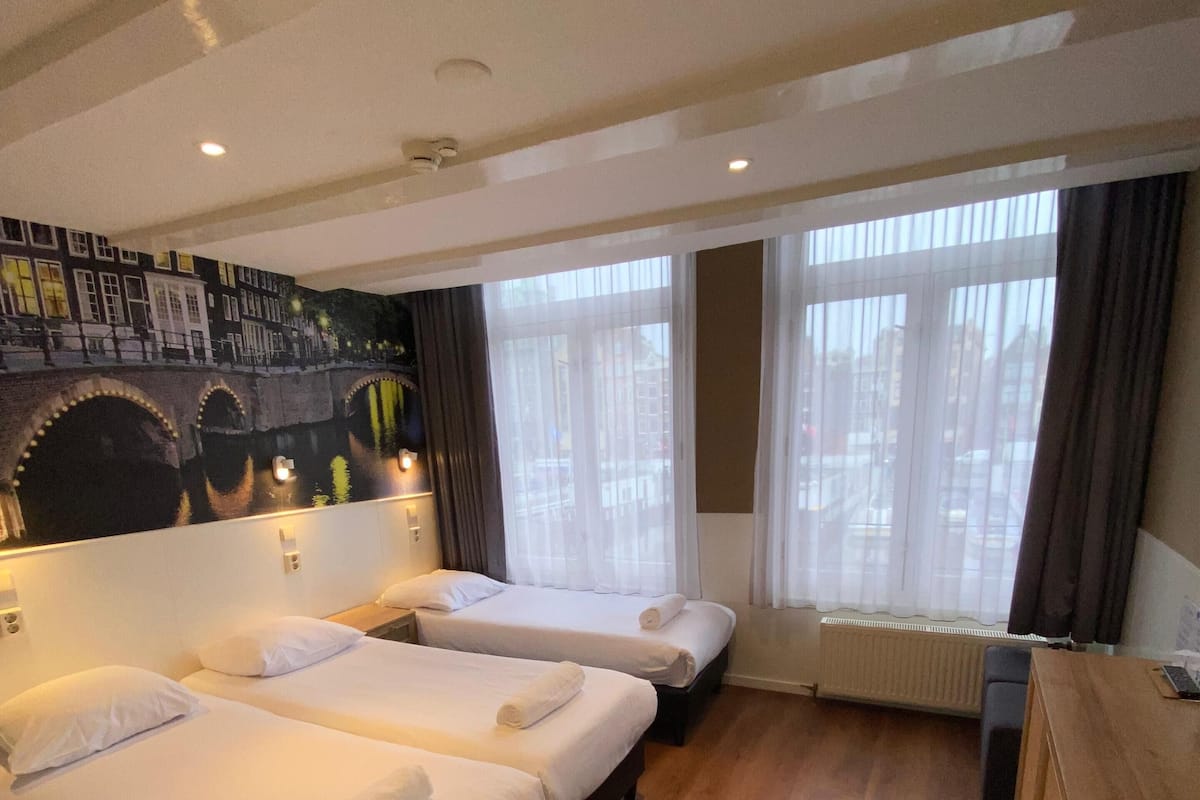A hotel room with three single beds, a wooden floor, a large window with curtains, and a mural of a bridge.