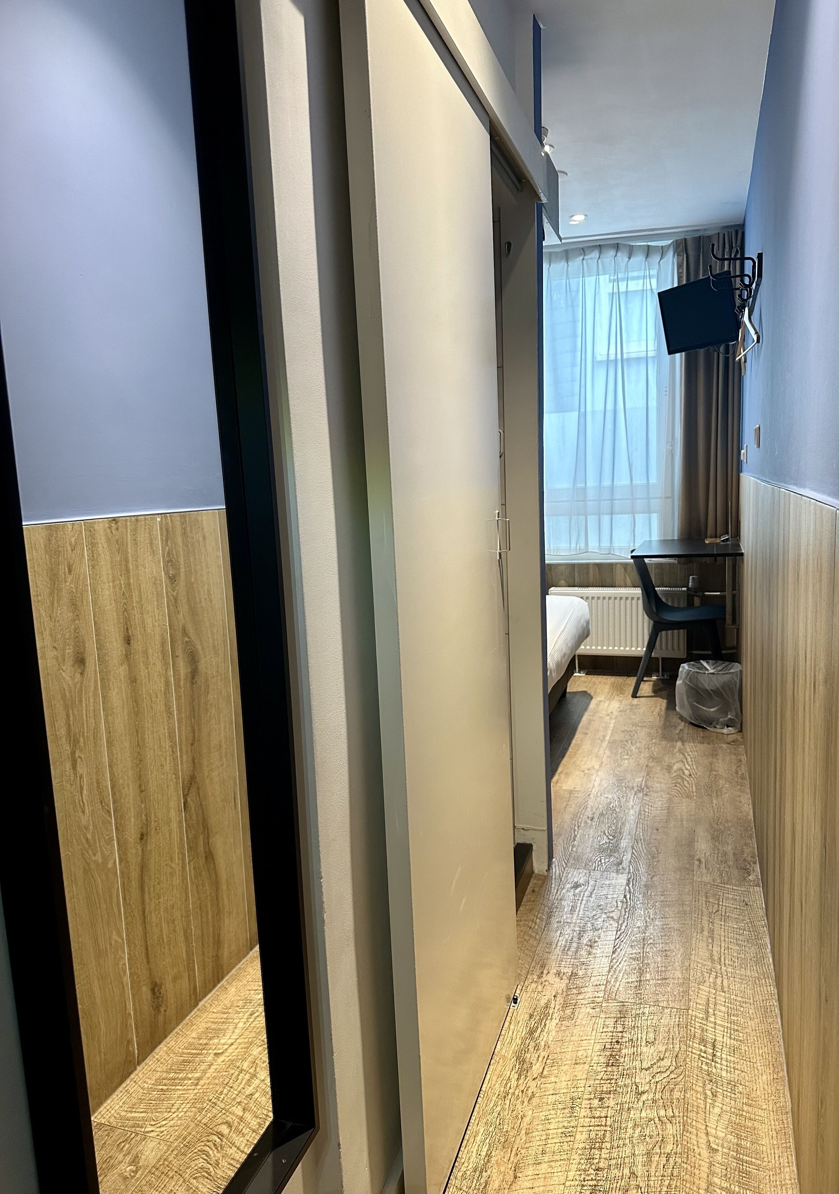 A hotel room with a wooden floor, a bed, a desk with a chair, and a TV mounted on the wall.