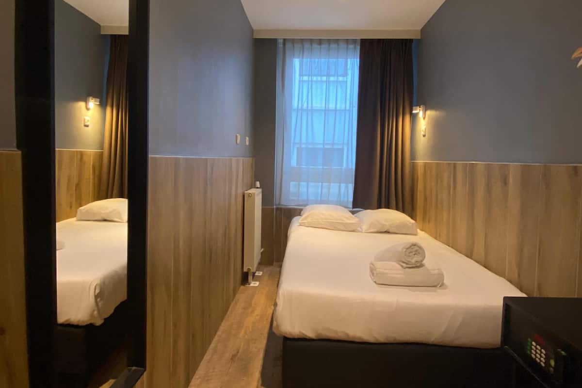A hotel room with a large mirror, a bed with white linens, a bedside table, and a window with curtains.