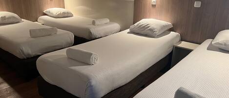 A hotel room with four single beds, each with a white mattress and pillow, and a wooden headboard.