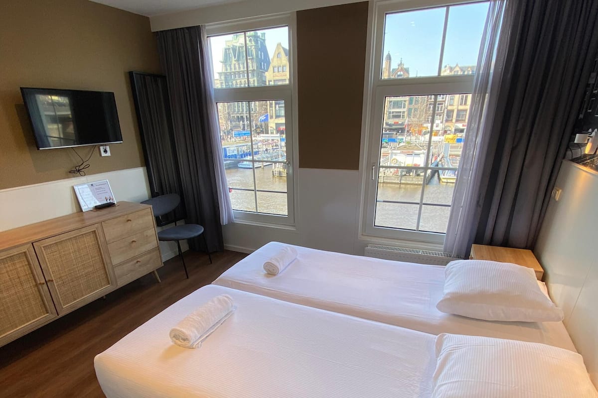 A hotel room with two beds, a TV, a desk, and large windows with a view of a cityscape.