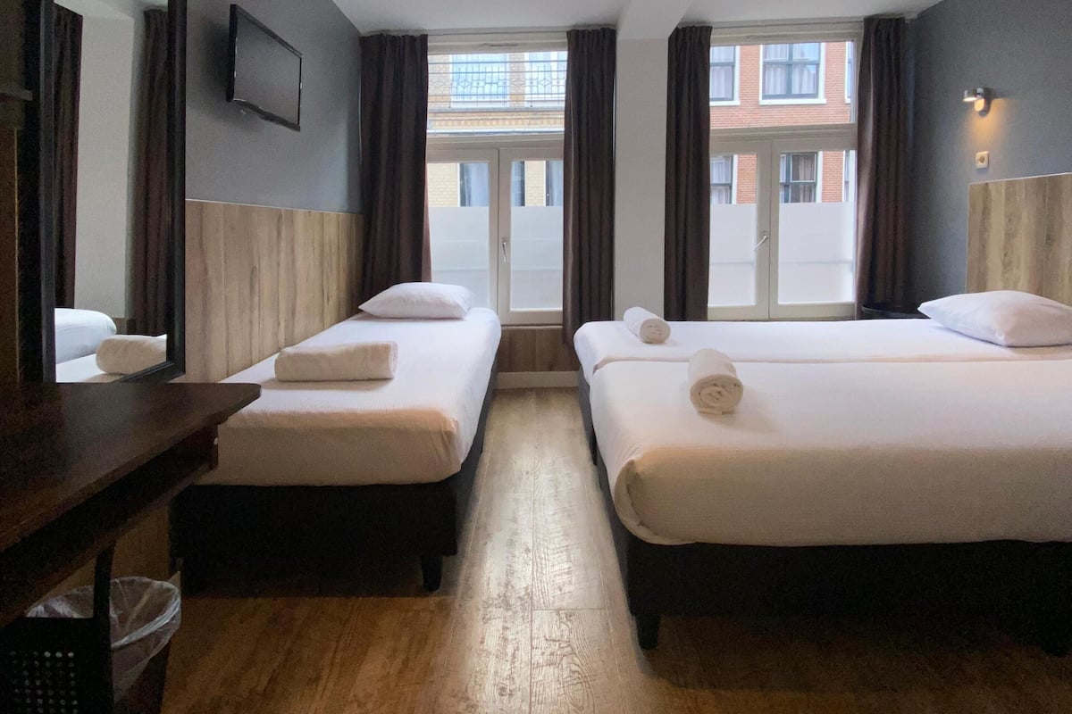 A hotel room with two single beds, a desk with a chair, a large mirror, and a view of the outside building through a window.