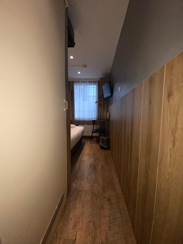A narrow hallway with a wooden floor leading to a bedroom with a bed, a window with curtains, and a mounted TV on the wall. - Hotel Old Quarter (Amsterdam)