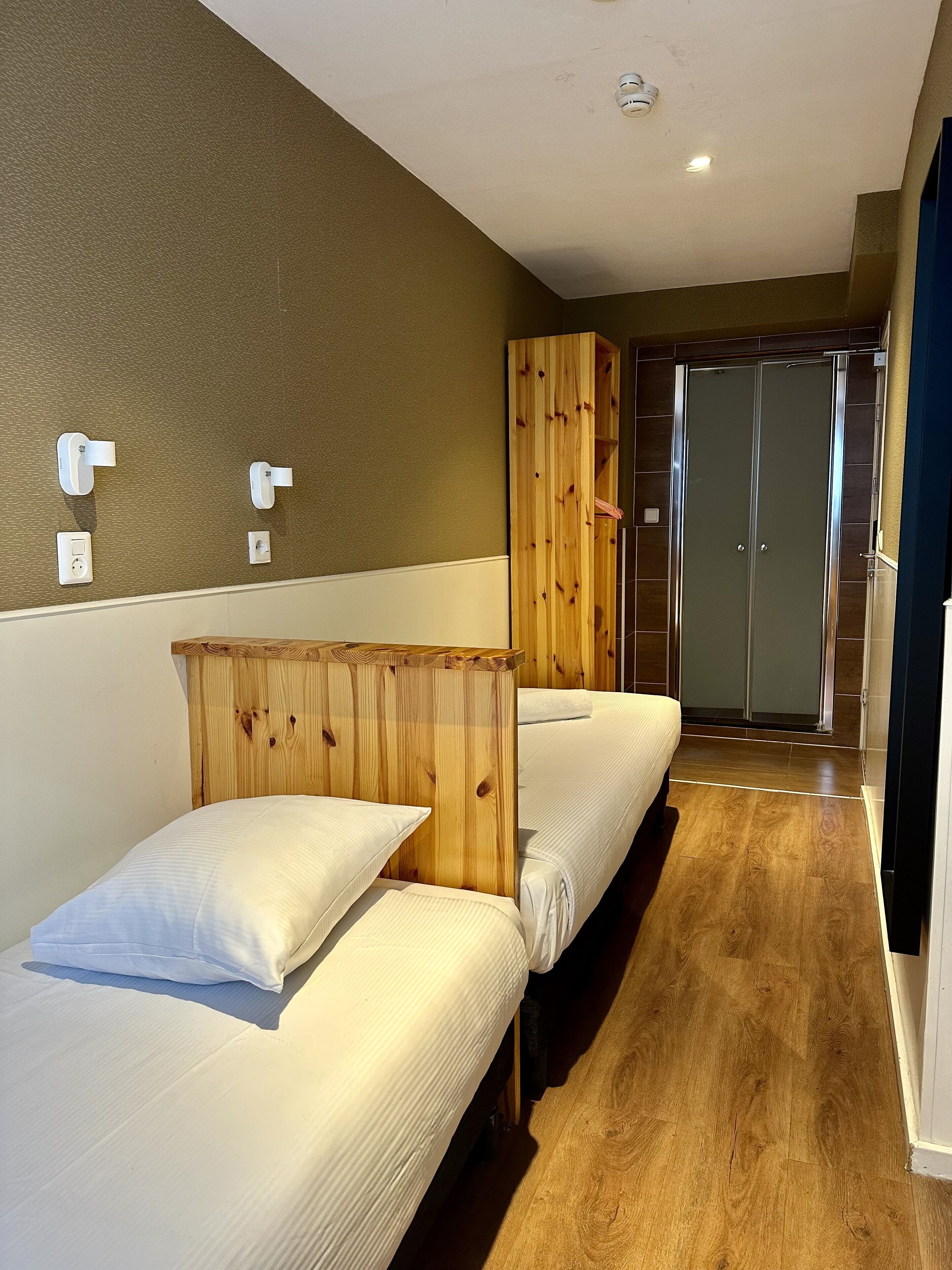 Habitación de hotel con dos camas individuales, un armario de madera, un baño con puerta de cristal y suelo de madera.