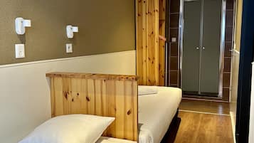 Habitación de hotel con dos camas individuales, un armario de madera, un baño con puerta de cristal y suelo de madera.