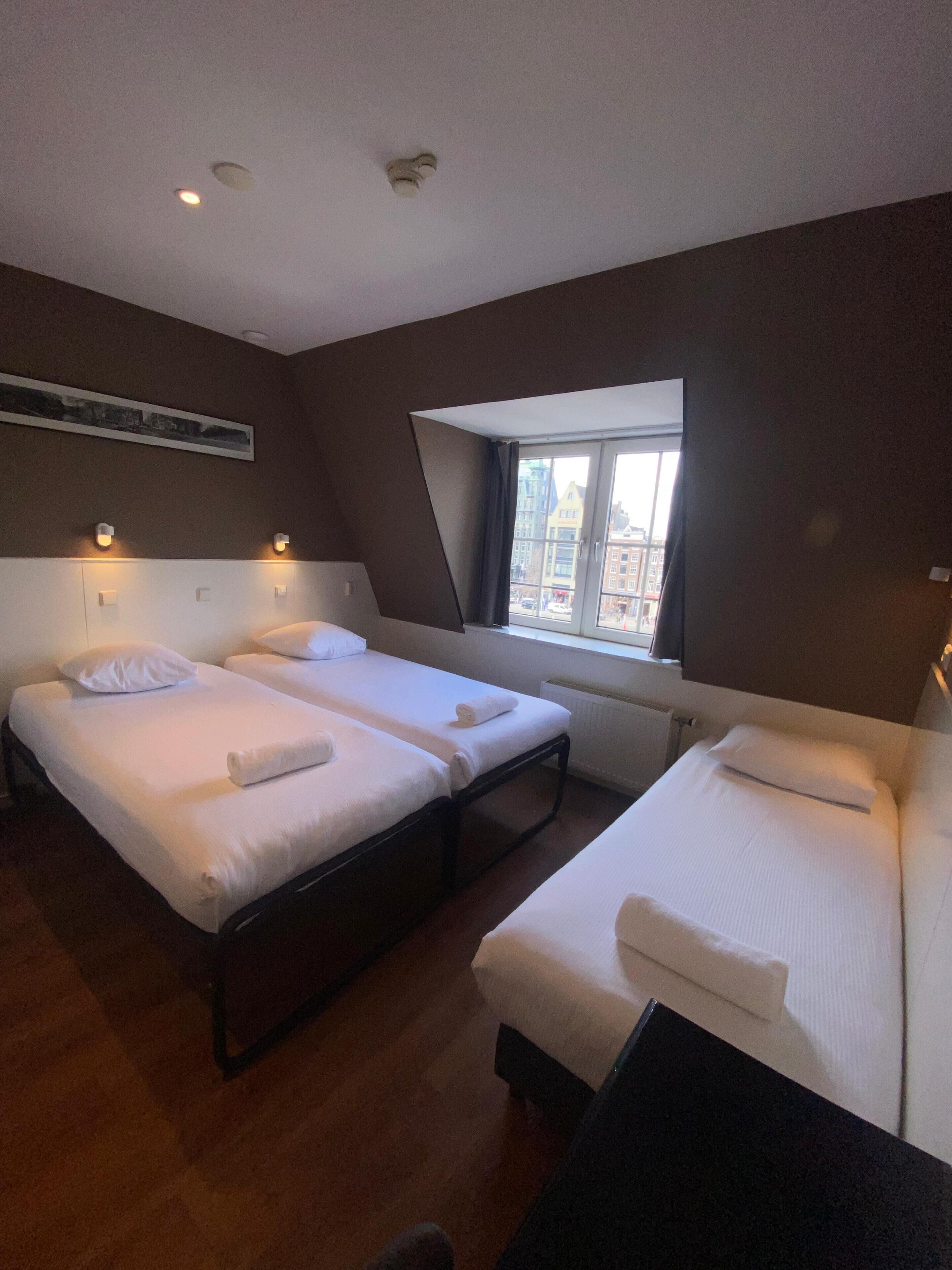 A hotel room with two beds, a window with a view of buildings, and a ceiling with recessed lighting.