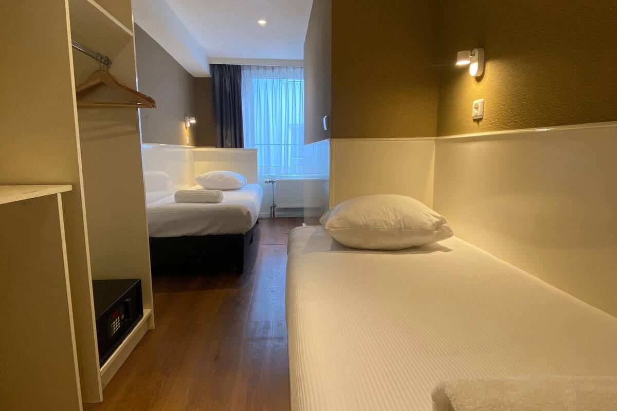 A hotel room with two beds, a wardrobe, a control panel, and a window with curtains.