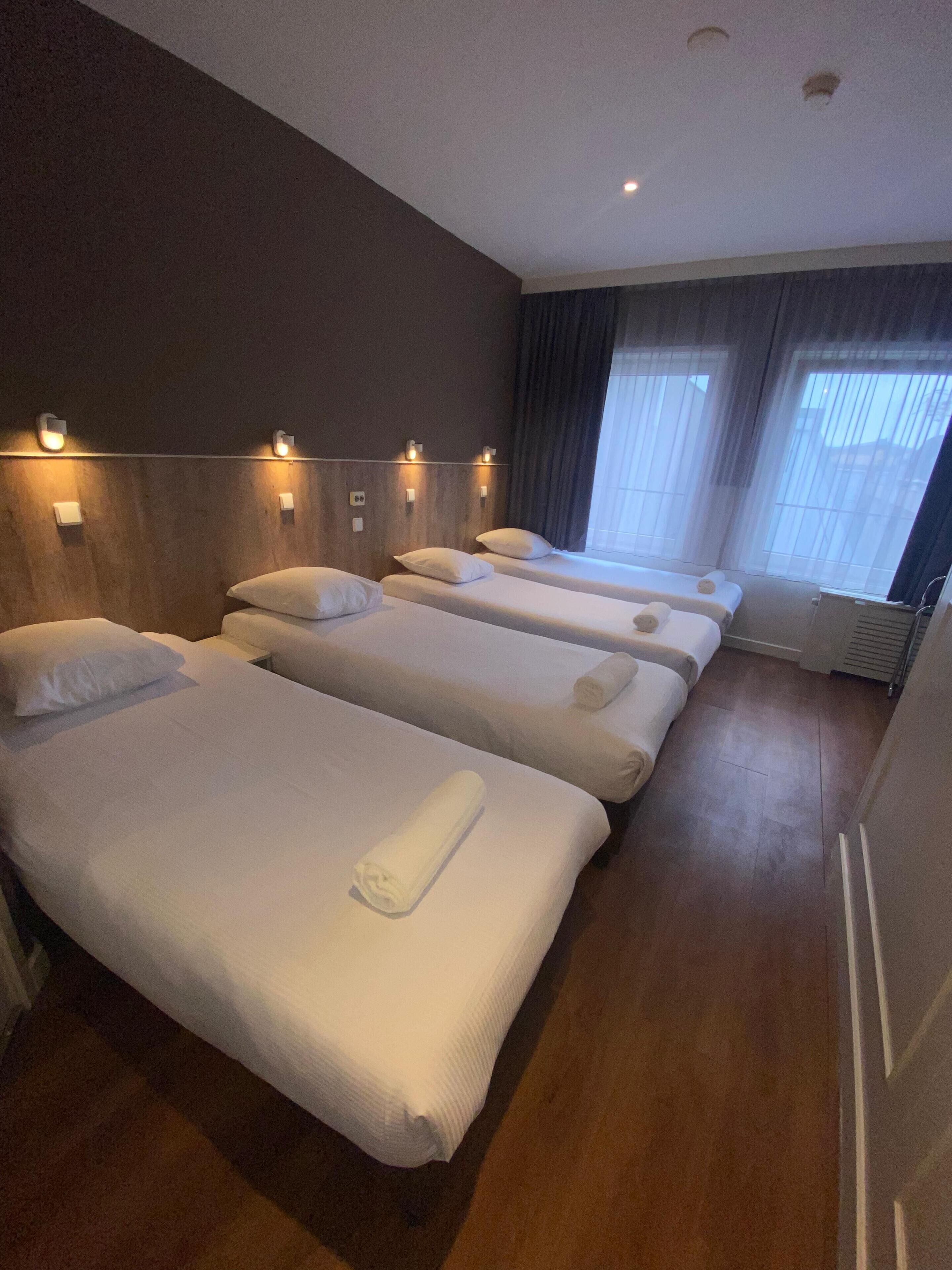 Habitación de hotel con cuatro camas individuales, cada una con una toalla enrollada, cabeceros de madera y luces fijadas a la pared.