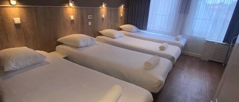 Habitación de hotel con cuatro camas individuales, cada una con una toalla enrollada, cabeceros de madera y luces fijadas a la pared.