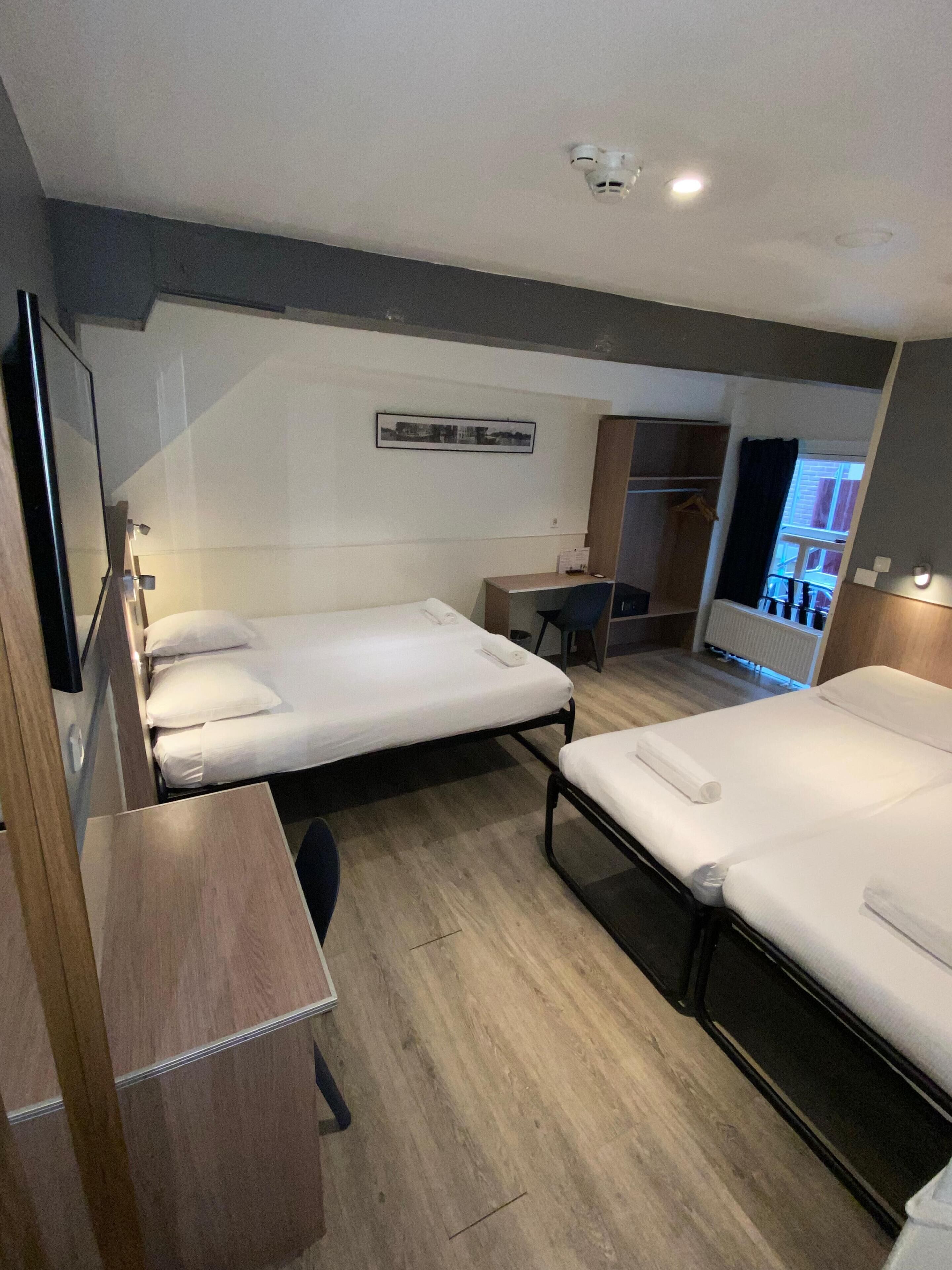 A hotel room with two single beds, a desk, a chair, and a small table. There is a wardrobe and a mirror on one wall, and a window with curtains.