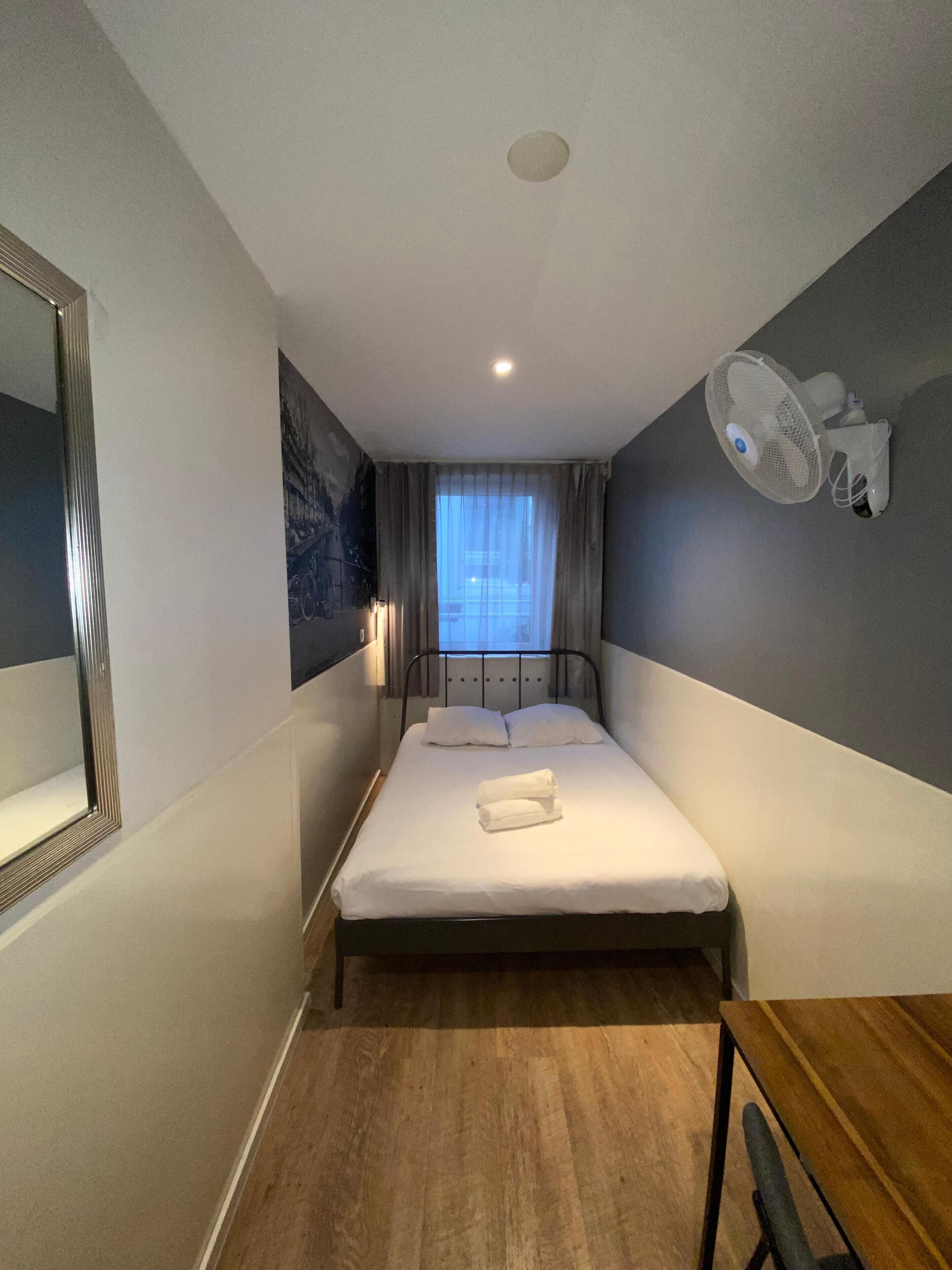 A small, neatly arranged hotel room with a single bed, a bedside table, a chair, a wall-mounted fan, and a window with curtains.