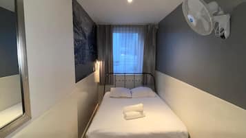 Ein kleines, ordentlich eingerichtetes Hotelzimmer mit einem Einzelbett, einem Nachttisch, einem Sessel und einem Wandventilator. Ein Fenster mit Vorhängen ist ebenfalls vorhanden.