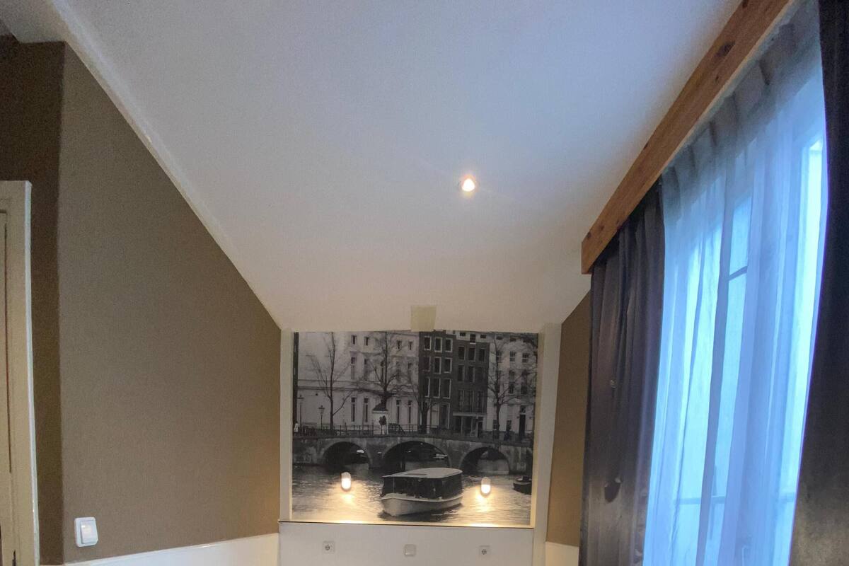 A hotel room with two single beds, a large window with curtains, a wall-mounted painting of a canal scene, and a wooden floor.