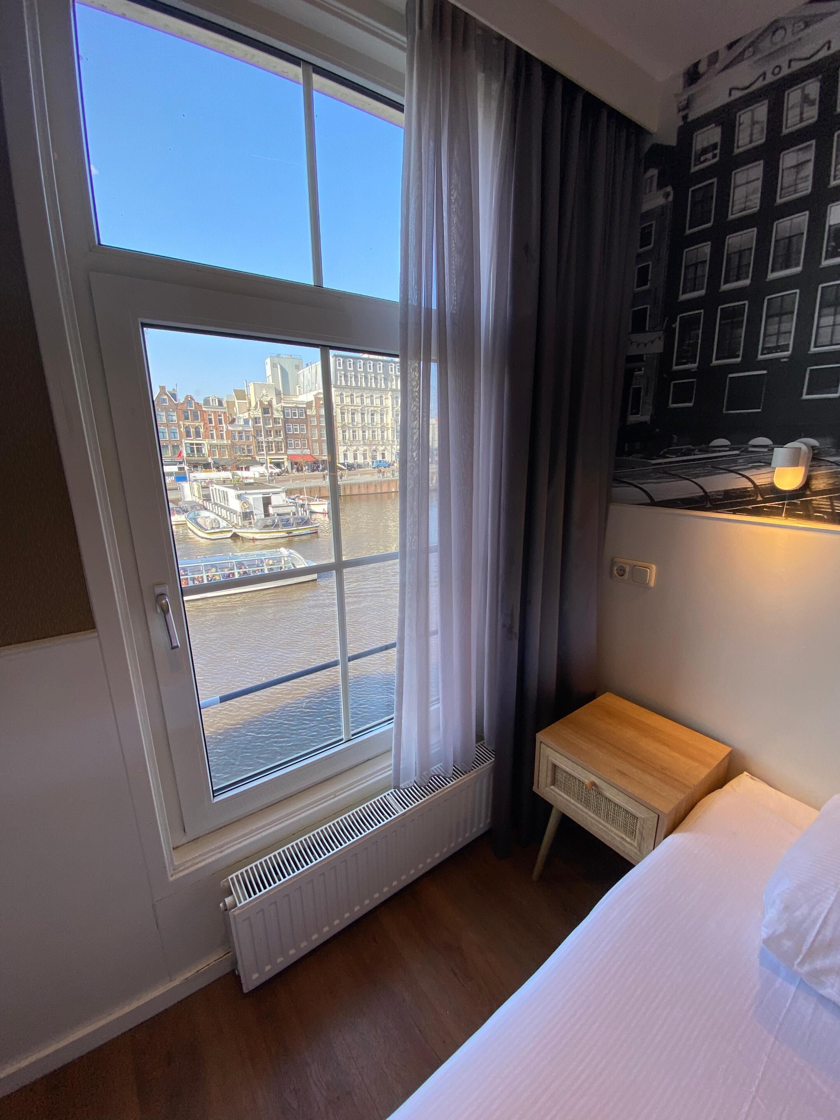 A hotel room with a large window overlooking a canal and historic buildings, a wooden bedside table, a radiator, and a wall with framed pictures.