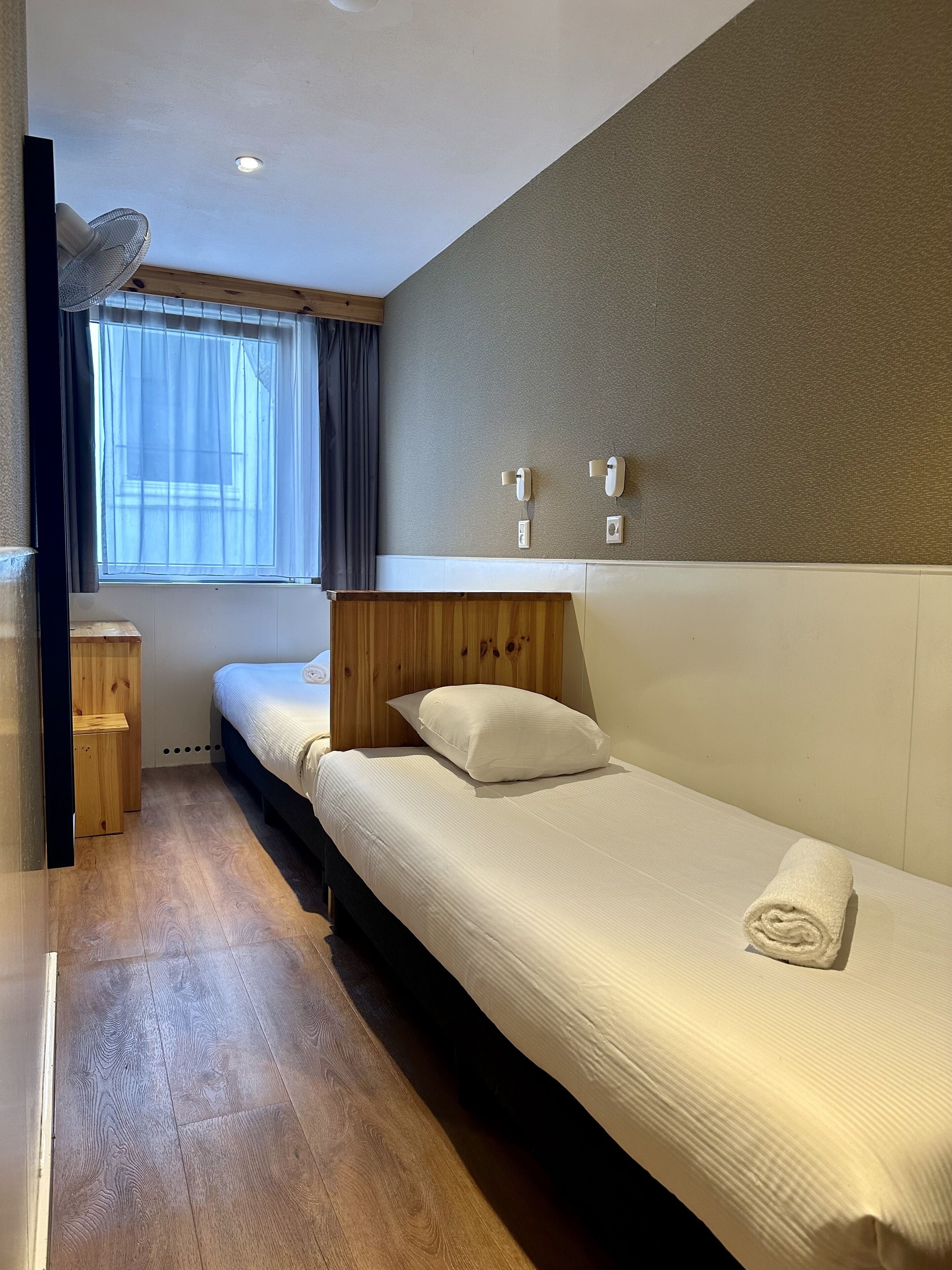 A hotel room with a wooden floor, a large bed with a white headboard, a smaller bed, a window with curtains, and a wall-mounted phone.