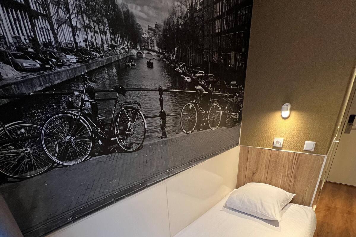 A hotel room with a single bed, a wall-mounted light, and a mural of a canal scene.