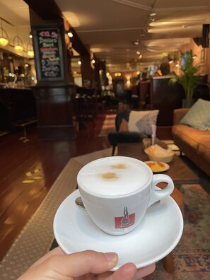 Una tazza di caffè su un piattino tenuto da una mano, con sullo sfondo l'interno di un caffè che include una poltrona, un tavolo con una ciotola e un menu scritto su una lavagna.