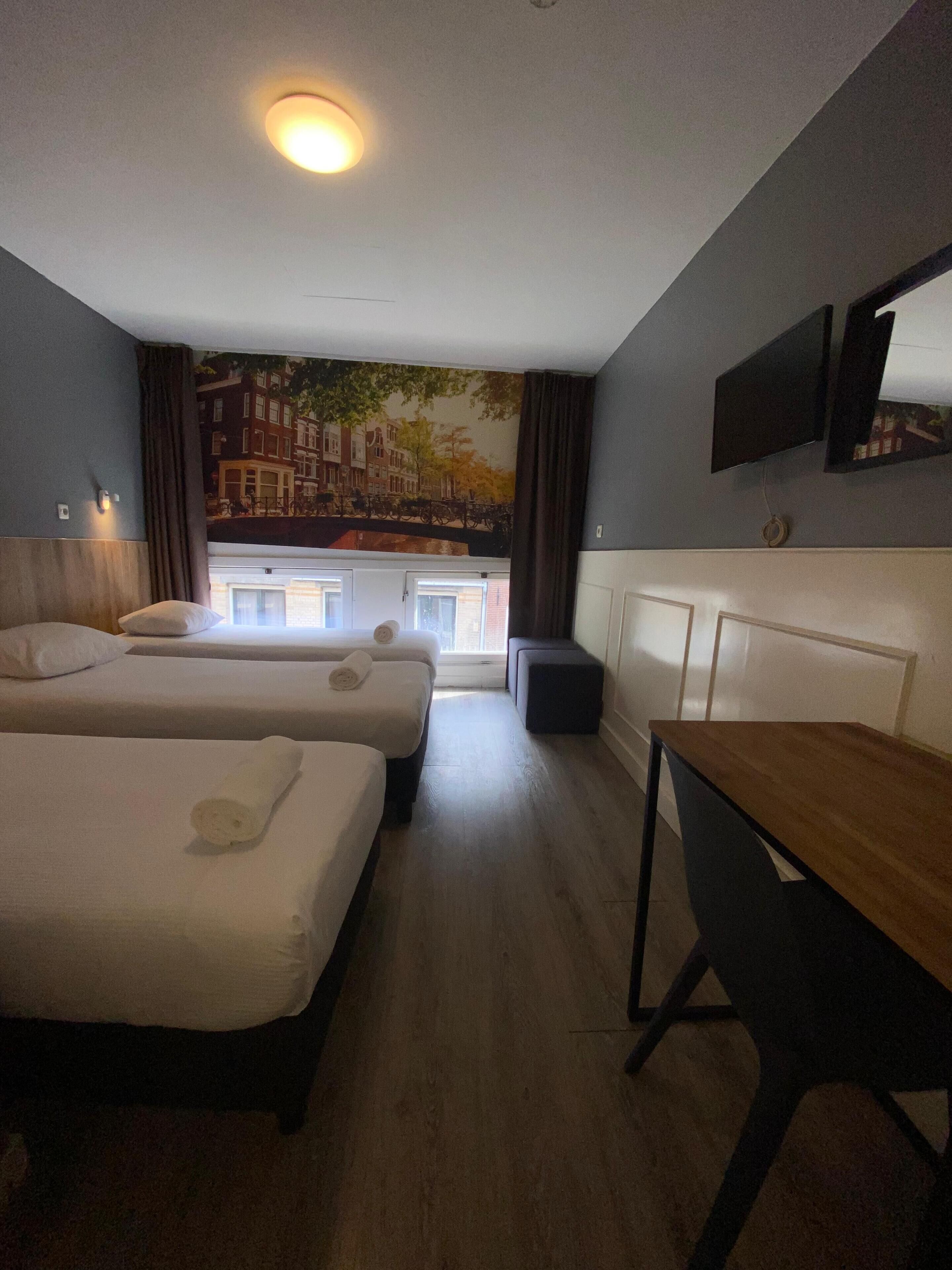 A hotel room with two beds, a desk, a chair, a TV, and a mural of a cityscape.
