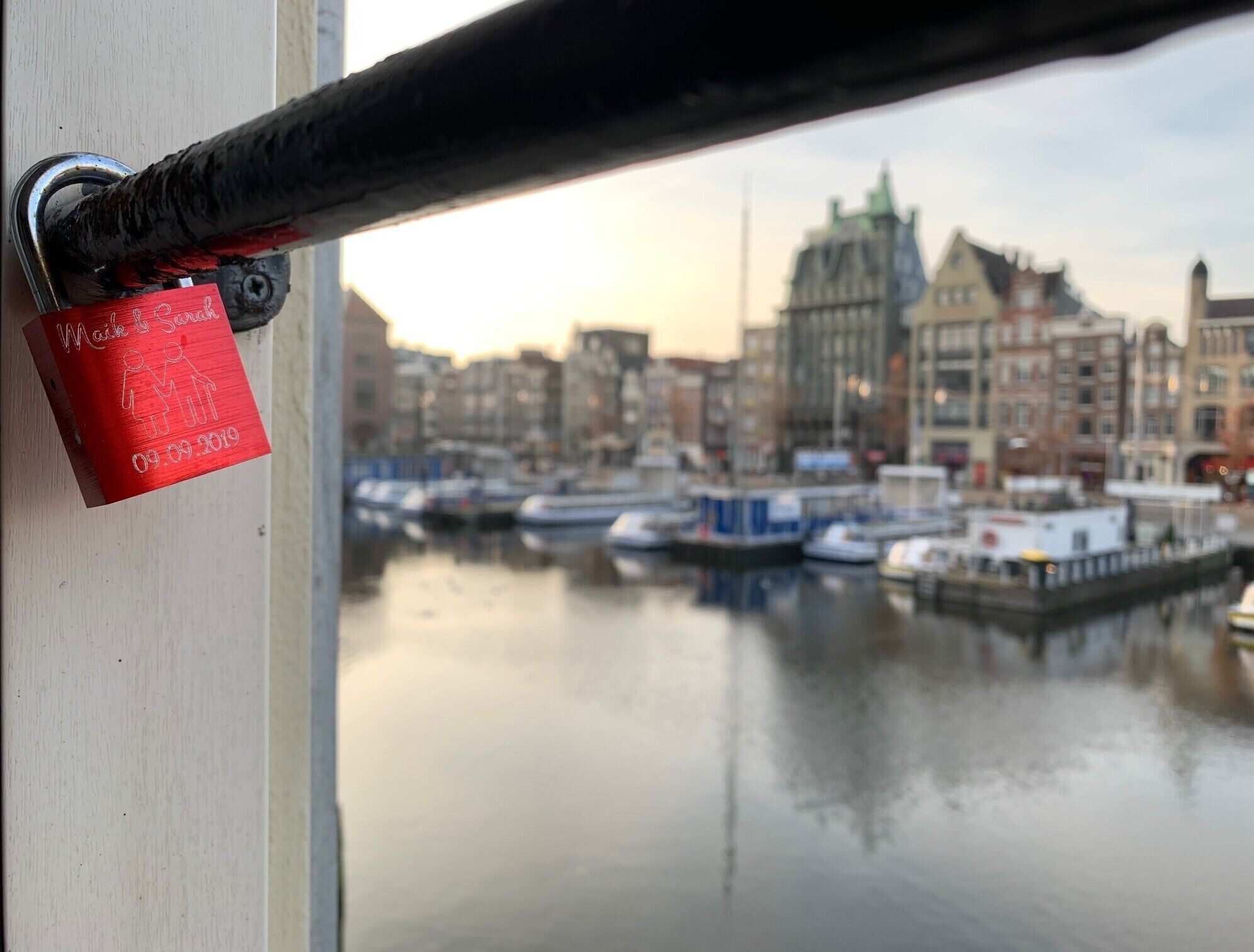 Un candado rojo con una fecha y nombres en una superficie metálica, con vista a una masa de agua tranquila con barcos y edificios al fondo.
