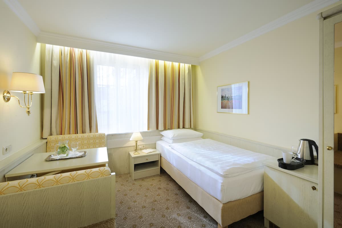 A hotel room with a bed, a small desk with a lamp, a chair, a nightstand, and a window with curtains.
