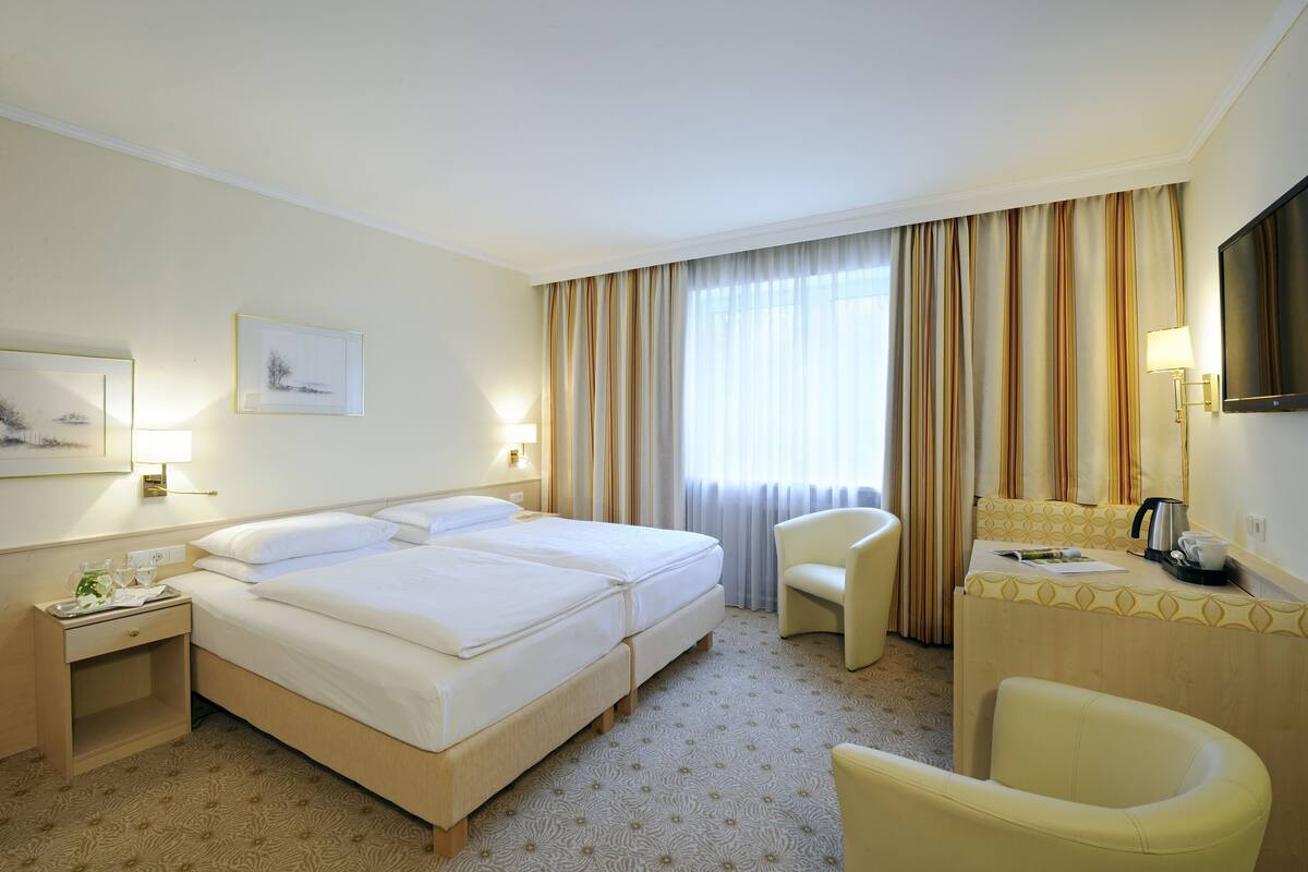 A hotel room with a double bed, a desk with a chair, a TV, and a window with curtains.