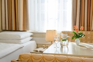 Una habitación de hotel con una cama, una mesita de noche con lámpara, un florero con flores y una ventana con cortinas.