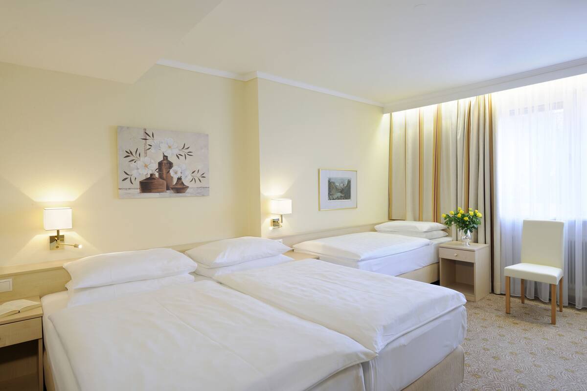 A hotel room with two beds, a nightstand with a lamp, a chair, a window with curtains, and artwork on the wall.