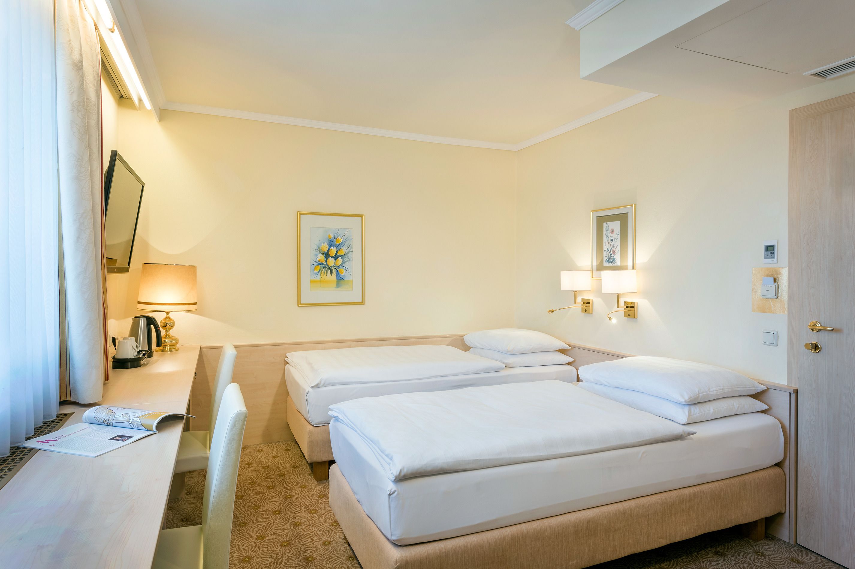 Habitación de hotel con dos camas, un escritorio con lámpara, un cuadro en la pared y una puerta.